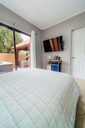 A bedroom with a large bed, a flat-screen TV mounted on the wall, a desk with a chair, and a sliding glass door leading to an outdoor area.