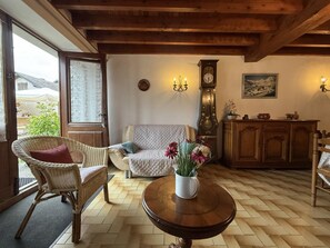 Ein gemütliches Wohnzimmer mit einem Korbstuhl, einer Couch, einem runden Couchtisch mit Blumen, einer Wanduhr und einem Gemälde.