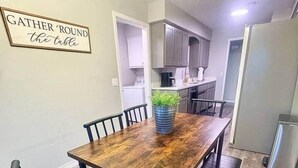Uma sala de jantar com uma mesa de madeira e cadeiras pretas, uma cozinha ao fundo e uma placa na parede.