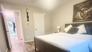 Quarto com cama, mesa de cabeceira, uma obra de arte na parede e porta que dá para outro cômodo.