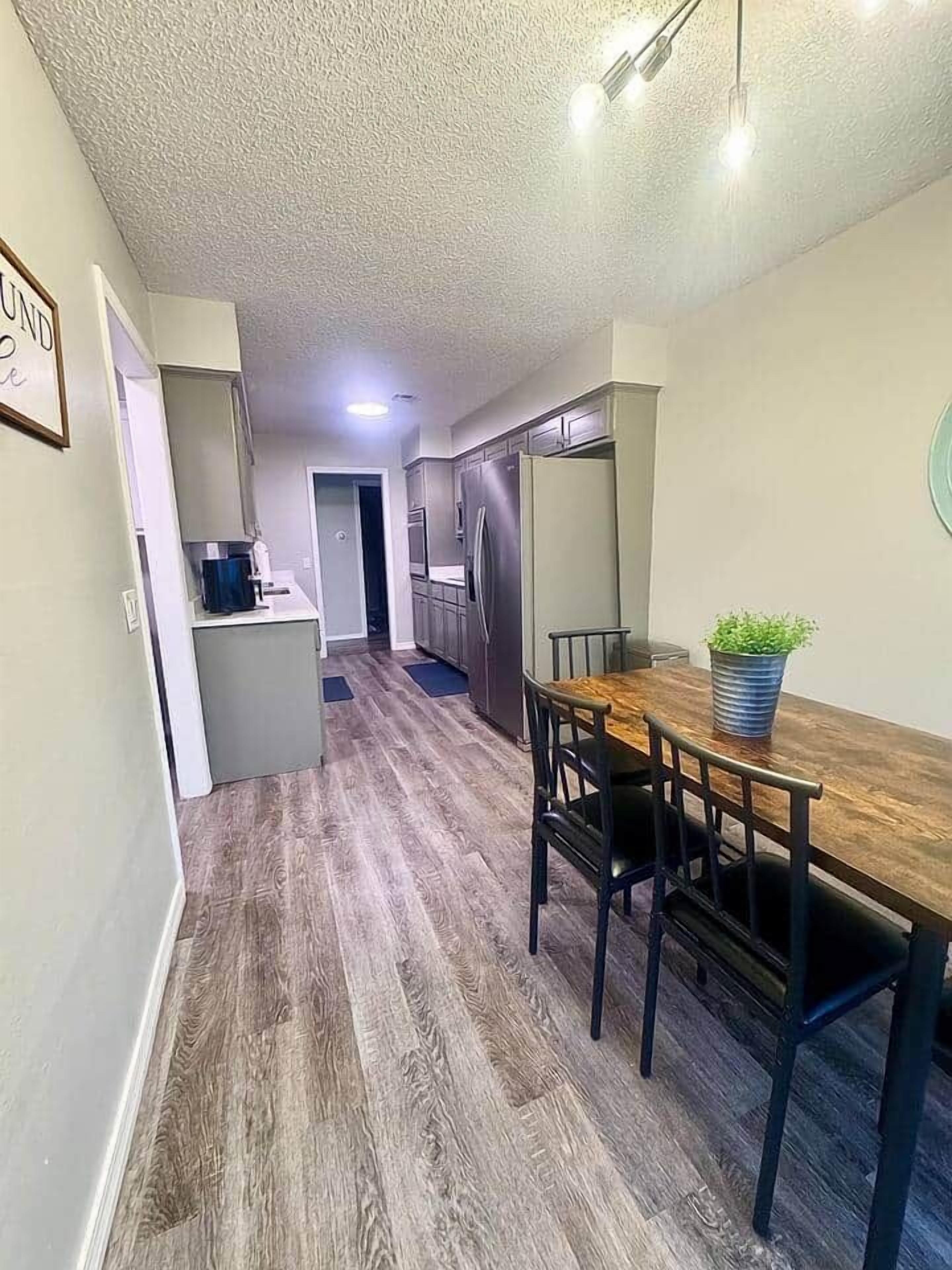 A narrow hallway leading to a dining area with a wooden table and chairs, a small kitchen area, and a door leading to another room.