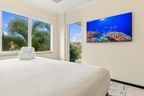 A hotel room with a bed, a window with a view of palm trees, and a wall-mounted TV displaying an underwater scene. - LeVent Sanctuary: Luxe Eagle Beach Condo 3BR 2BA (Eagle Beach)