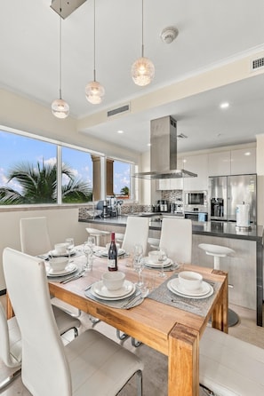 A modern kitchen with a dining area, large windows, and a view of palm trees. - LeVent Sanctuary: Luxe Eagle Beach Condo 3BR 2BA (Eagle Beach)