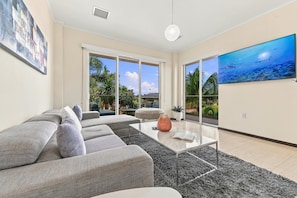 A modern living room with a grey sofa, a glass coffee table, and a large painting of an underwater scene. - LeVent Sanctuary: Luxe Eagle Beach Condo 3BR 2BA (Eagle Beach)