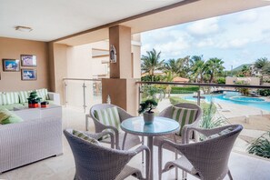 Ein Balkon mit Korbmöbeln, einem Glastisch und Blick auf ein Schwimmbad und Palmen.