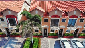 A row of multi-story buildings with red-tiled roofs, balconies, and a palm tree in the foreground. - Gold Coast Retreat: 2BR/3BA Townhome, Private Pool (Noord)