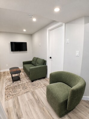 A living room with a sofa, a chair, a small table, and a flat-screen TV mounted on the wall. - Vine Luxe.

A brand-new, stylish and peaceful private 2-bedroom. (Regina)