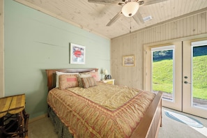 Ein Schlafzimmer mit einem Bett, einem Holz-Kopfteil, einem Deckenventilator und einer Tür zu einer grünen Fläche.