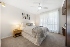 A bedroom with a bed, bedside lamps, a ceiling fan, a window with sheer curtains, and a nightstand. - Broadstairs—Spacious Beachside Garden Escape (Port Elliot)