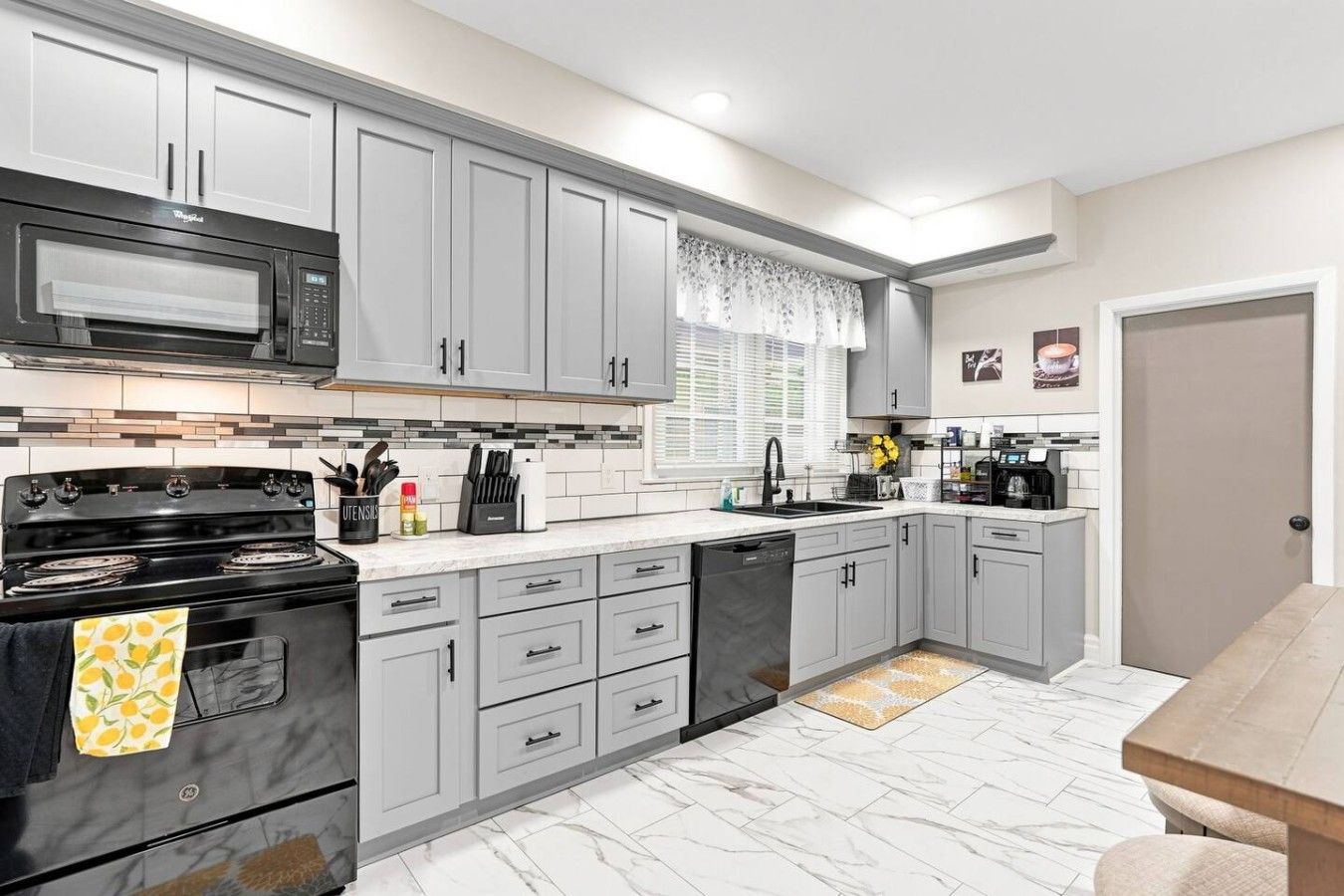 A modern kitchen with grey cabinets, a black stove, and a microwave. There is a window with a white curtain, a sink, and a coffee maker.