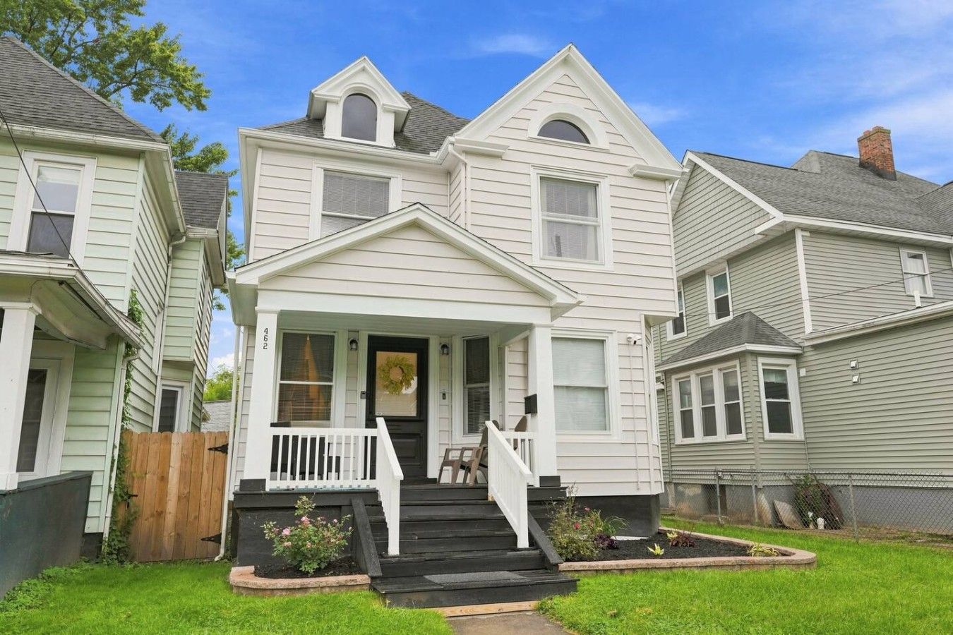 A two-story house with a front porch, a visible front door, and a small front yard with a wooden fence and some plants.
