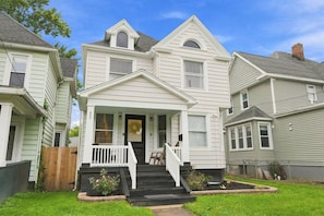 A two-story house with a front porch, a visible front door, and a small front yard with a wooden fence and some plants. - Beautifully Remodeled 5BR Home Near Niagara Falls! (Niagara Falls)