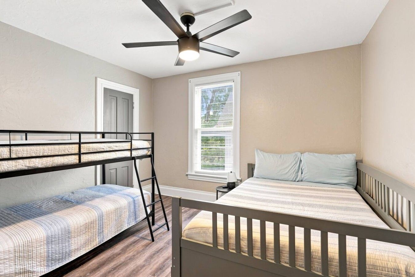 A bedroom with bunk beds, a ceiling fan, a window with blinds, and a bedside table.