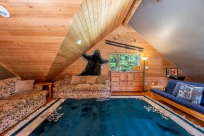 A cozy attic room with a sloped ceiling, a large rug, a sofa, a cabinet, and a window with a view of trees. - Mountain Escape. Sauna & Soak. (Columbia-Shuswap)