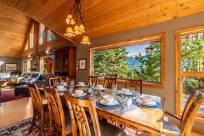 A dining area with a wooden table set for six, surrounded by chairs, with large windows offering a view of trees and mountains. - Mountain Escape. Sauna & Soak. (Columbia-Shuswap)