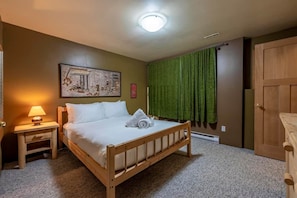 A hotel room with a wooden bed, a nightstand with a lamp, a painting on the wall, and a green curtain. - Mountain Escape. Sauna & Soak. (Columbia-Shuswap)