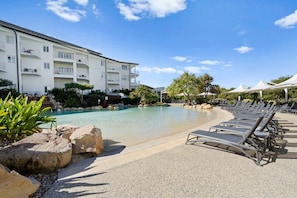 Ein Schwimmbecken mit Liegestühlen, ein Gebäude mit Balkonen und ein klarer Himmel.