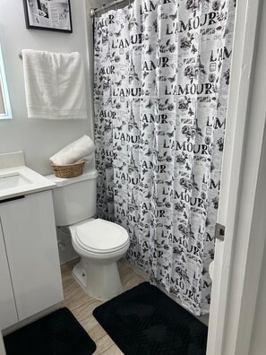 Un baño con inodoro, un lavamanos blanco, una toalla blanca, un felpudo negro y una cortina de ducha estampada.