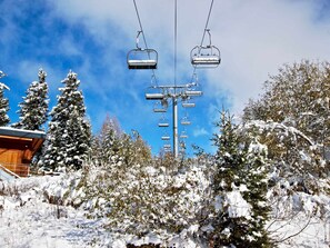 Eine Sesselbahn mit mehreren Sesseln, die an Seilen hängen, umgeben von schneebedeckten Bäumen und blauem Himmel.
