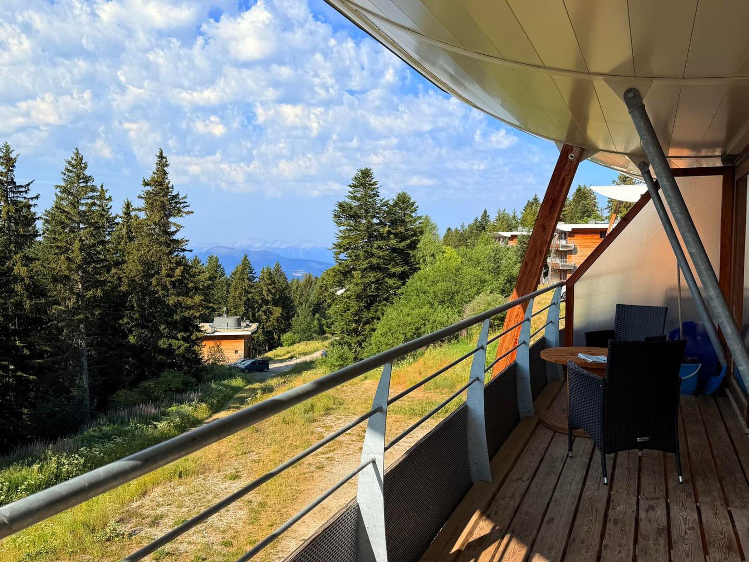 A balcony with a view of a forest and mountains, a wooden deck, and a covered seating area.