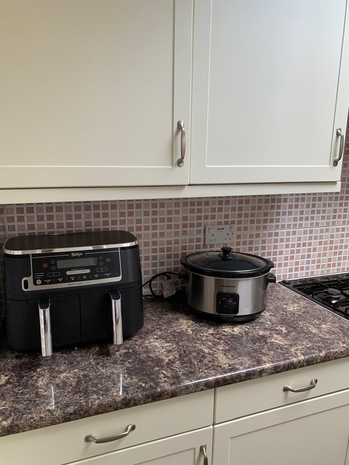 A kitchen with white cabinets, a black Ninja smart cook system, and a stainless steel slow cooker on a granite countertop.