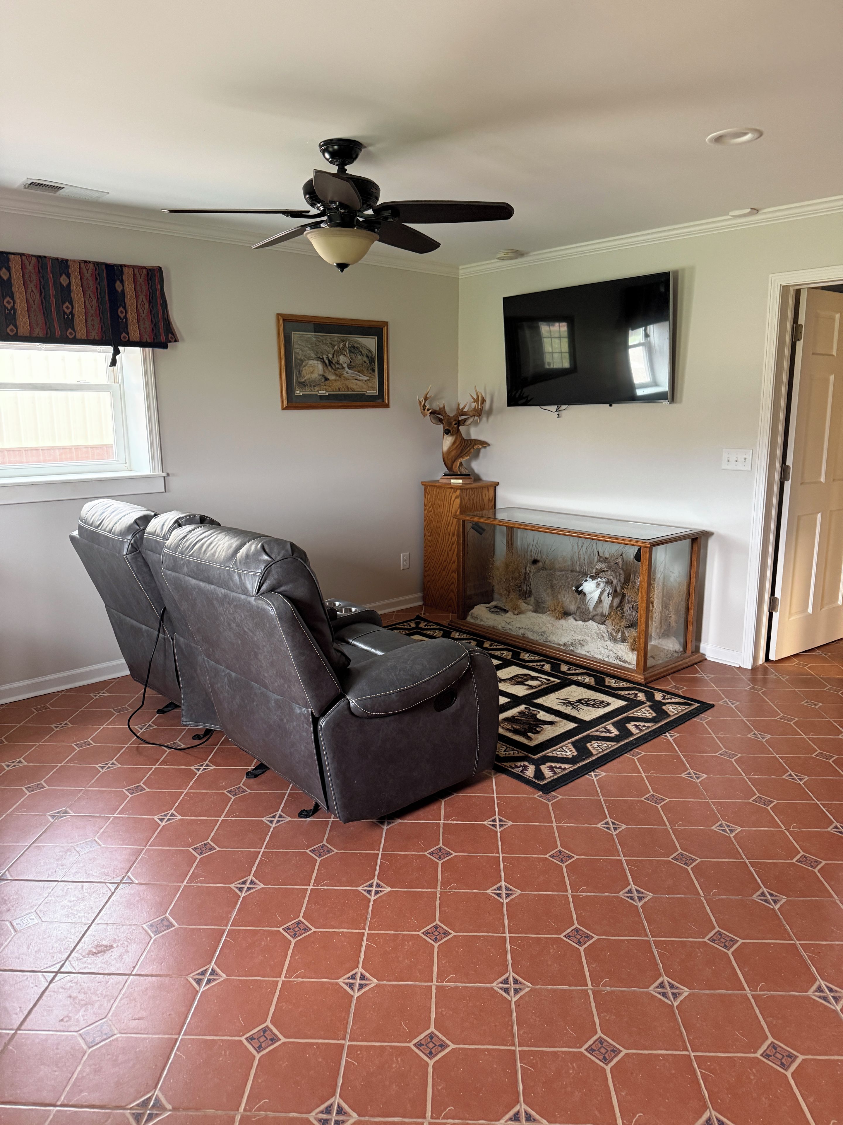 Un salon comprenant un fauteuil inclinable en cuir, un ventilateur de plafond, une cheminée ornée d’une tête d’animal décorative, un téléviseur fixé au mur et des carreaux de sol à motifs.