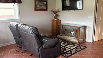 A living room with a leather recliner, a ceiling fan, a fireplace with a decorative animal head, a television mounted on the wall, and patterned floor tiles.