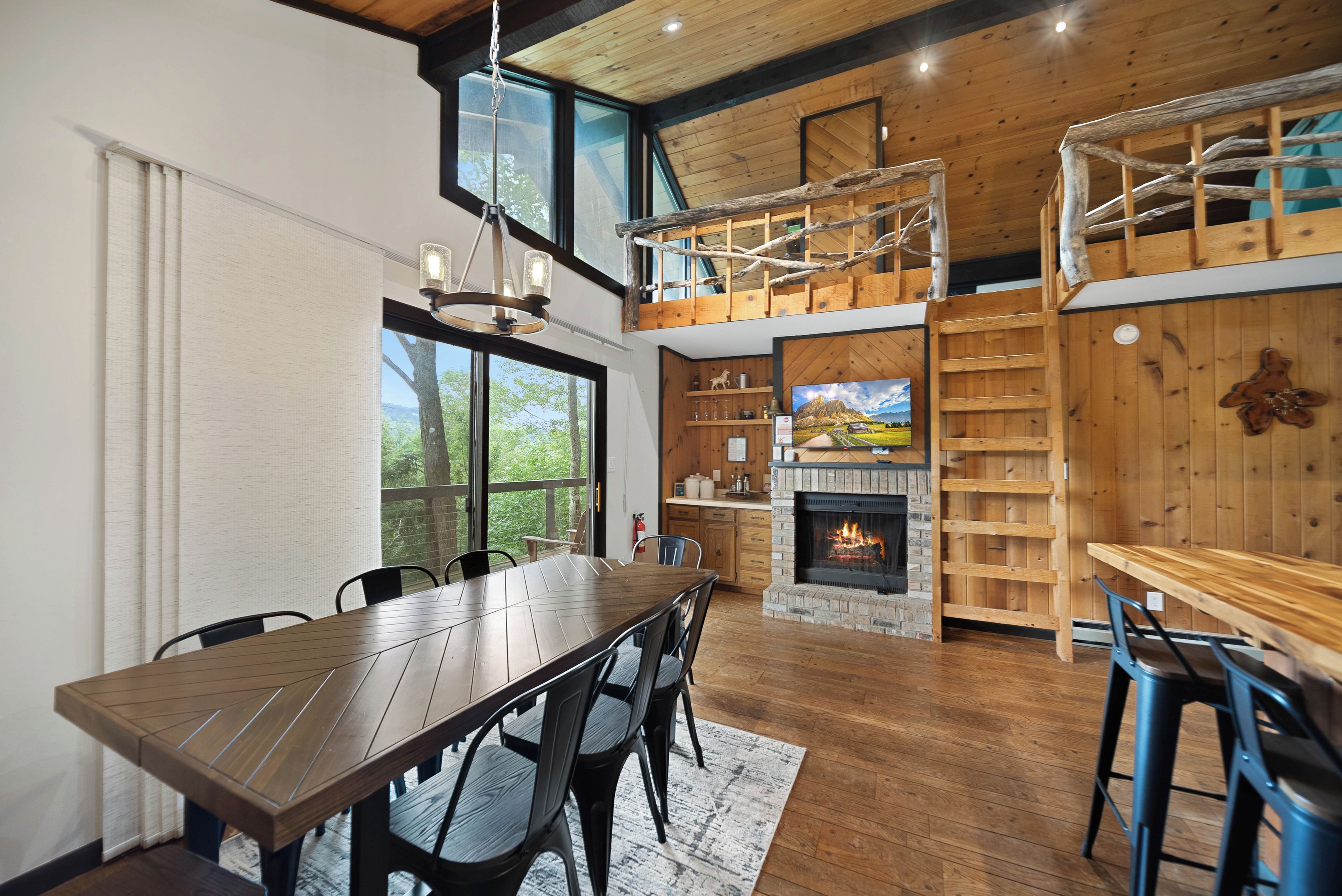 A spacious dining area with a long wooden table and chairs, a fireplace, and a loft with a wooden staircase.