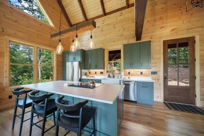 A modern kitchen with a central island, bar stools, and a view of the outdoors through large windows. - Charming Mountain Retreat Near Banner Elk (Banner Elk)