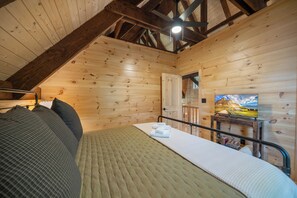 A wooden cabin bedroom with a sloped ceiling, a bed with pillows, a bedside table with a lamp, and a TV on the wall. - Charming Mountain Retreat Near Banner Elk (Banner Elk)