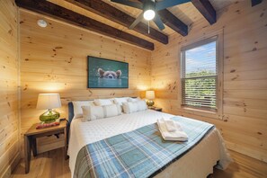 Ein Zimmer einer Holzhütte mit einem Bett, zwei Nachttischlampen, einem Fenster mit Jalousien und einem gerahmten Bild an der Wand.
