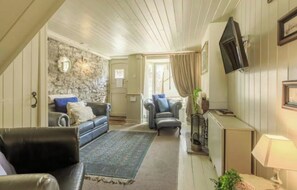 Un salon chaleureux avec un mur en pierre, un canapé en cuir noir, un fauteuil bleu et un téléviseur fixé au mur.