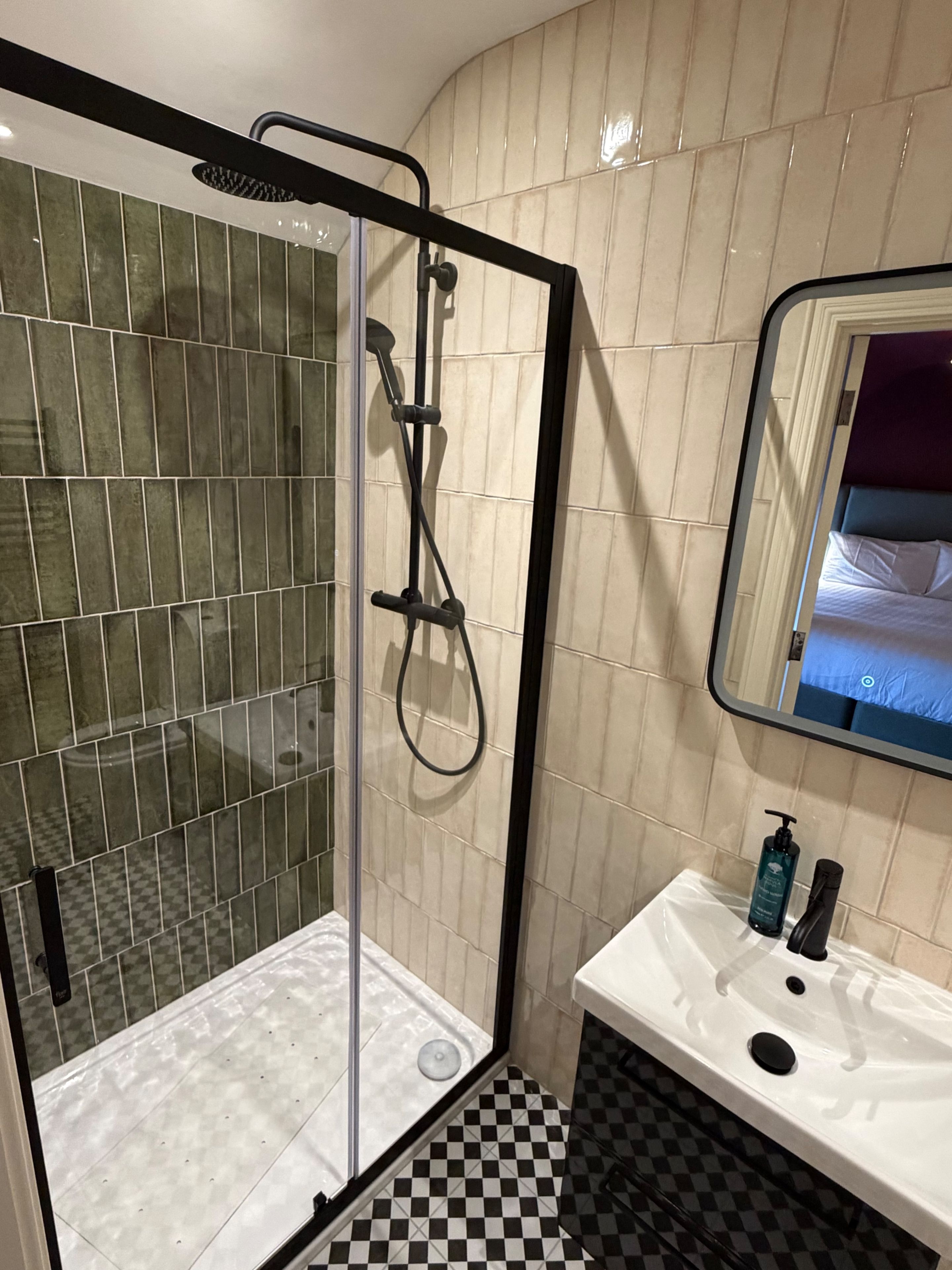Une salle de bain avec une douche en verre, un lavabo blanc et un sol à carreaux noirs et blancs, ainsi qu’un miroir qui reflète un lit.