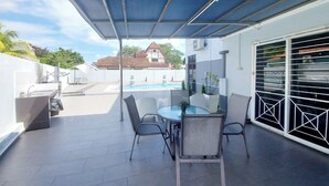 An outdoor patio with a covered seating area, a pool in the background, and a white building with a garage. - Melaka A’Famosa Grand Pool Villa – Private Pool & Family Getaway (Alor Gajah)