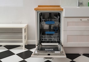 Een open vaatwasser in een keuken met een geruit vloertje, een witte tafel en een wit kastje.