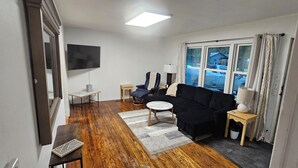 Una sala de estar con un sofá negro, una pequeña mesa redonda, un televisor y un ventanal con cortinas.