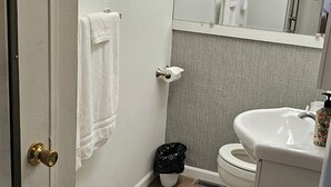 Un baño con inodoro, lavamanos y espejo. Hay toallas colgadas en la pared y un cubo de basura.