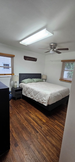 Un dormitorio con cama, ventilador de techo, mesita de noche, lámpara y ventana con persianas.
