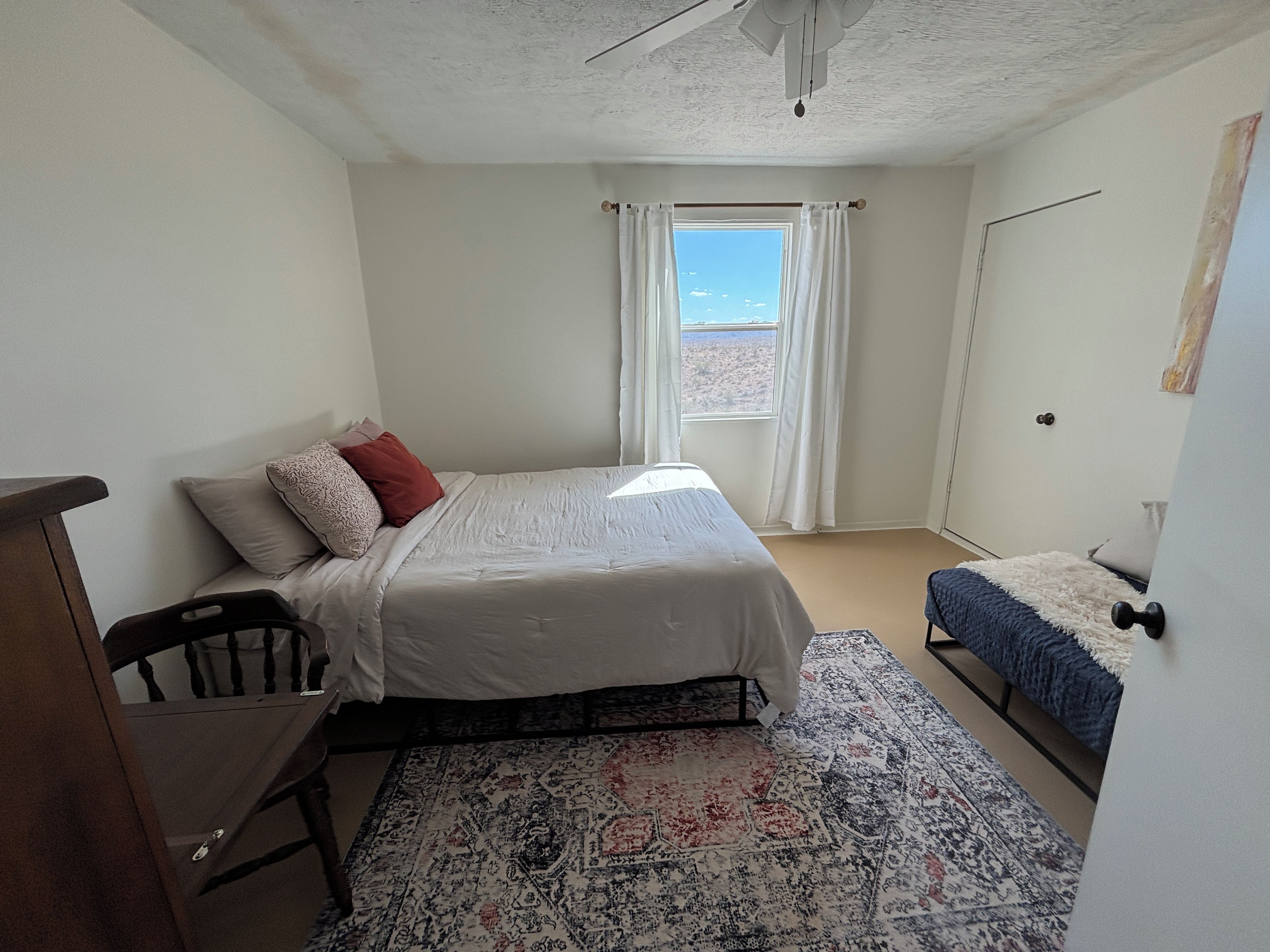 Un dormitorio con cama, escritorio, silla, ventana con cortinas y puerta.