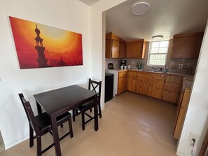 A dining area with a table and chairs, a kitchen with wooden cabinets and a window, and a painting on the wall. - Remote Alamo Lake Getaway!  Dark night skies. AZPT. Fishing. Farm. Ghost town. (Yucca)
