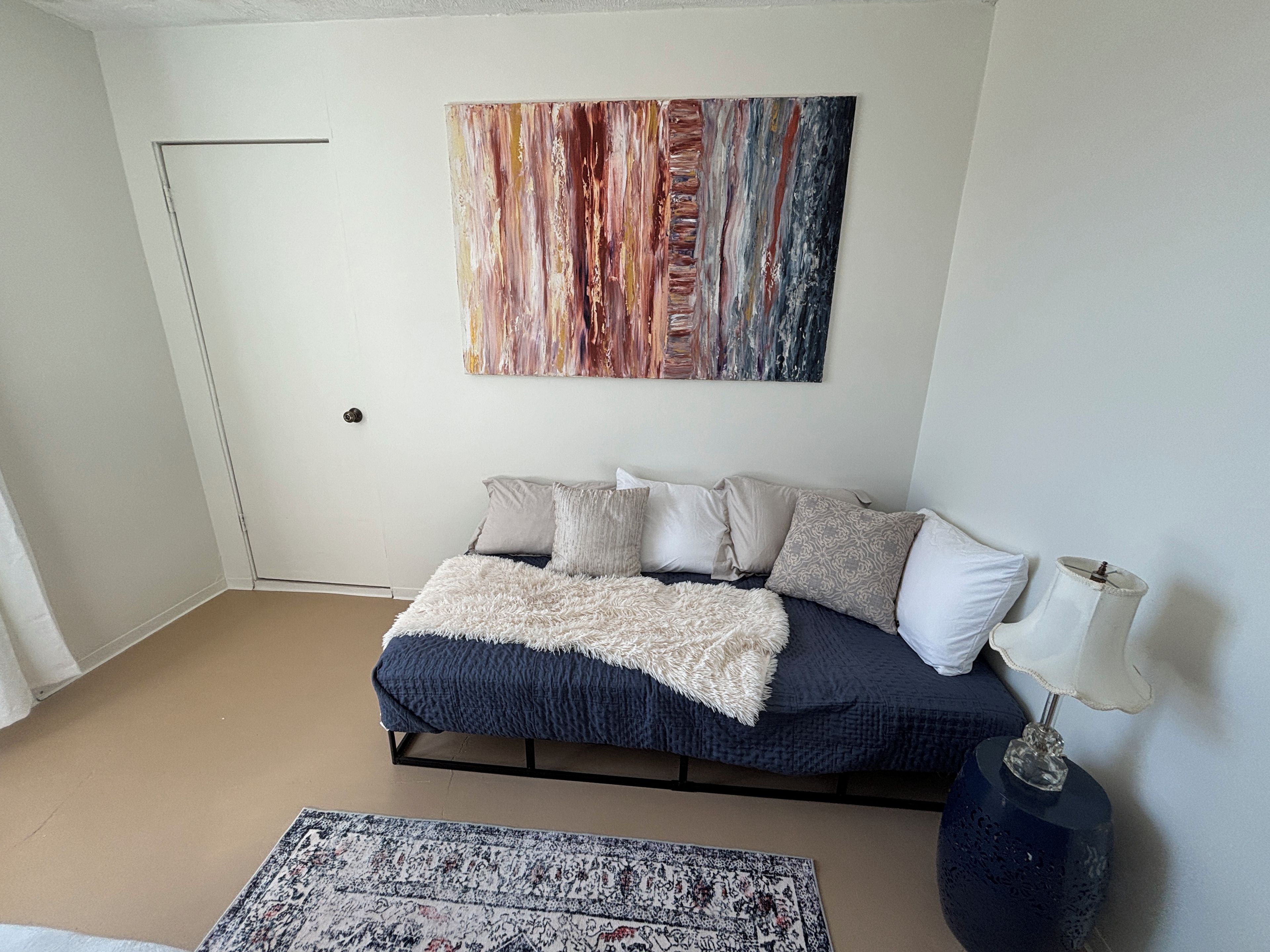 Un dormitorio con cama, almohadas, una colcha, un tapete, una obra de arte en la pared y una lámpara de noche.