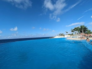 A clear blue pool with a view of the ocean and a beach area with palm trees and lounge chairs. - New stylish,1 BR, Resort access, SOV - Sea-Esta (Sosua)