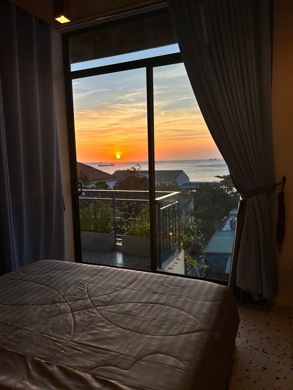 A bedroom with a large window overlooking a sunset, a bed, and a view of a balcony with plants.