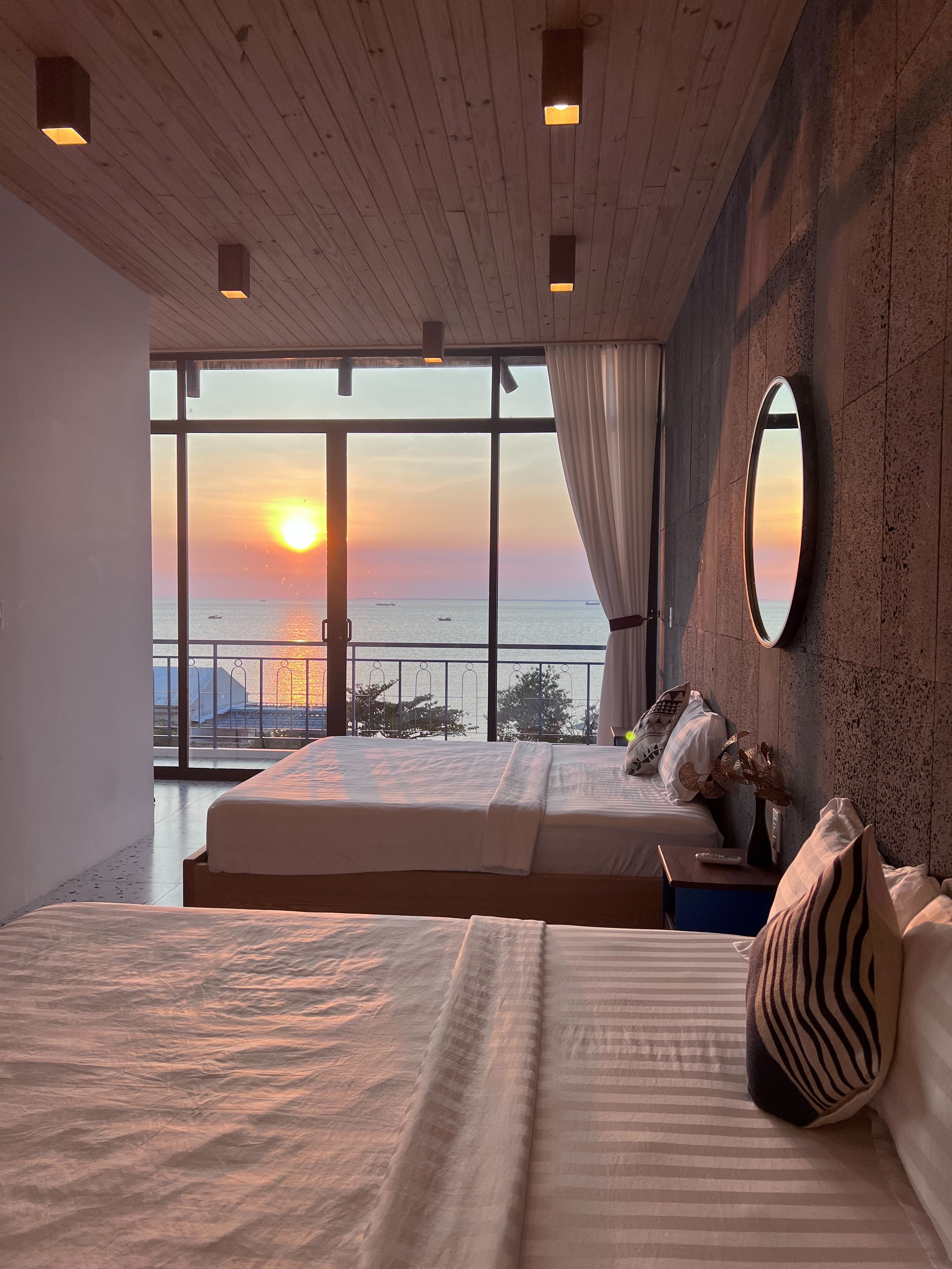 Ein Hotelzimmer mit einem großen Bett, einem Nachttisch, einem Spiegel und Blick auf den Sonnenuntergang über dem Meer.