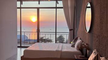 Ein Hotelzimmer mit einem großen Bett, einem Nachttisch, einem Spiegel und Blick auf den Sonnenuntergang über dem Meer.