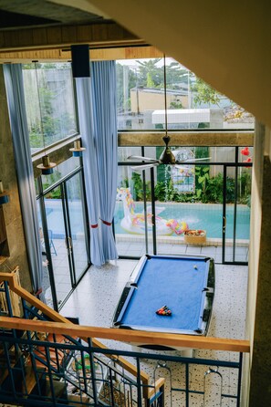 Un tavolo da biliardo in una stanza con ampie finestre, una ventola a soffitto e vista su una piscina esterna.
