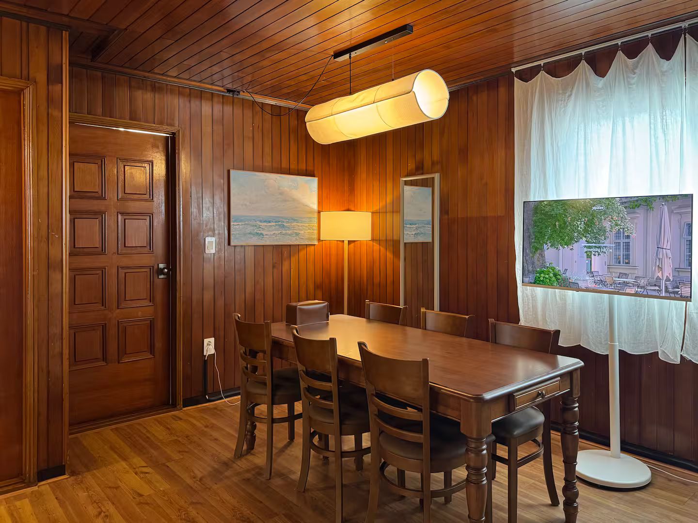 A wooden-paneled room with a dining table, chairs, a lamp, and a window with a view of a building.