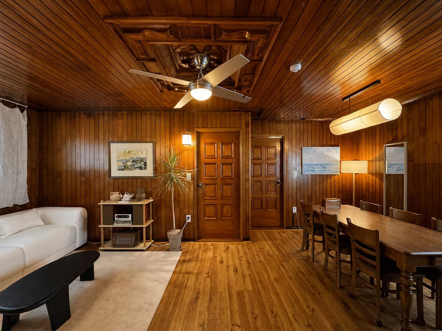 A room with wooden paneling, a ceiling fan, a dining table with chairs, a white sofa, and a small table with a plant and a lamp.