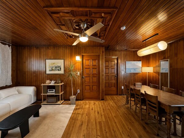 A room with wooden paneling, a ceiling fan, a dining table with chairs, a white sofa, and a small table with a plant and a lamp.