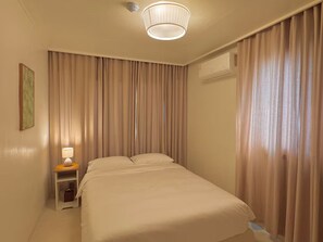 A hotel room with a bed, a bedside table with a lamp, curtains, and an air conditioning unit.